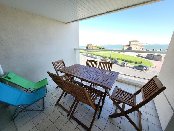 Un balcon avec une table et des chaises en bois, une chaise pliante verte, et une vue sur une route et des immeubles.