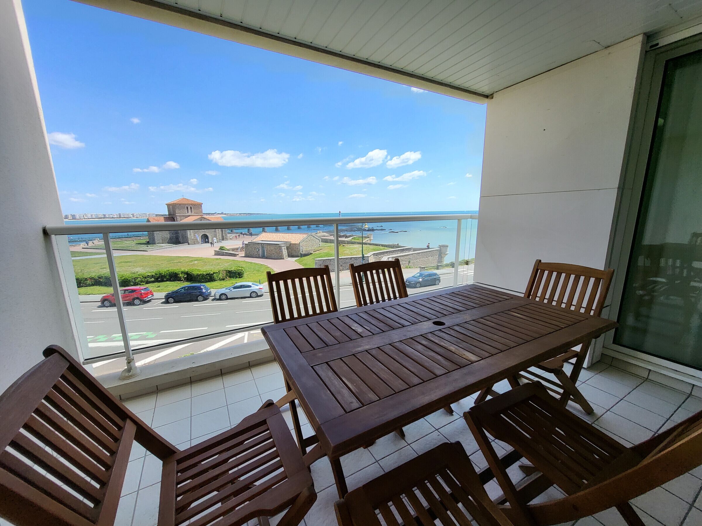 En balkon med et træbord og stole, udsigt over et kystområde med bygninger og biler.