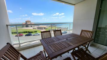 Un balcon avec une table et des chaises en bois, donnant sur une zone côtière avec des bâtiments et des voitures.