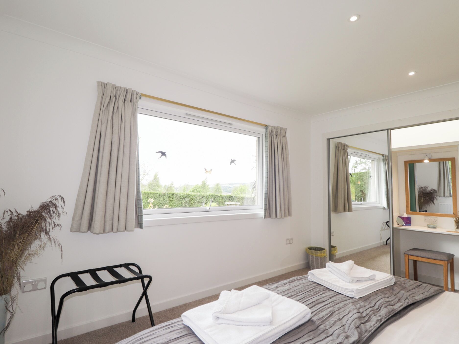 A modern bedroom with a large window, a bed with neatly folded towels, a bedside table, a mirror, and a potted plant.