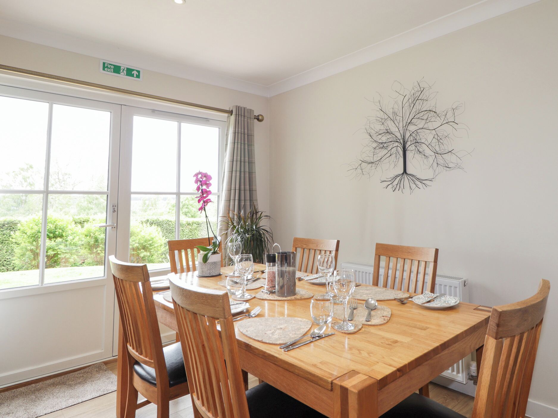 A dining room with a wooden table set for four, chairs, a potted plant, and a fire exit sign above a window.