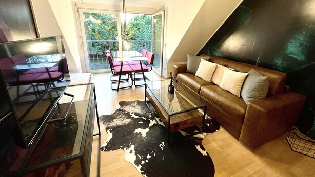A modern living room with a glass coffee table, leather sofa, and a dining area with a glass table and chairs. - Boutique Apartment Petite Belle (Freiburg im Breisgau)