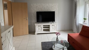 Een woonkamer met een televisie op een witte kast, een rode bank, een glazen salontafel en een deur naar een andere kamer.