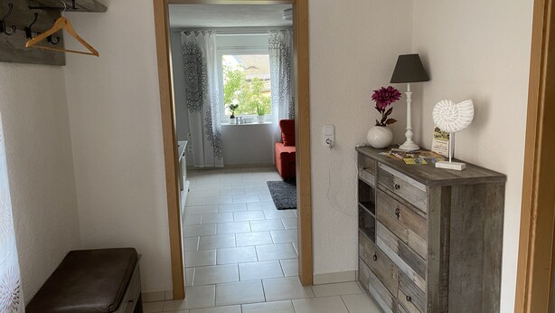 Een hal met een houten bankje, een dressoir met een lamp en decoratieve objecten, en een deur die leidt naar een kamer met een rode stoel en een raam met gordijnen met een patroon.