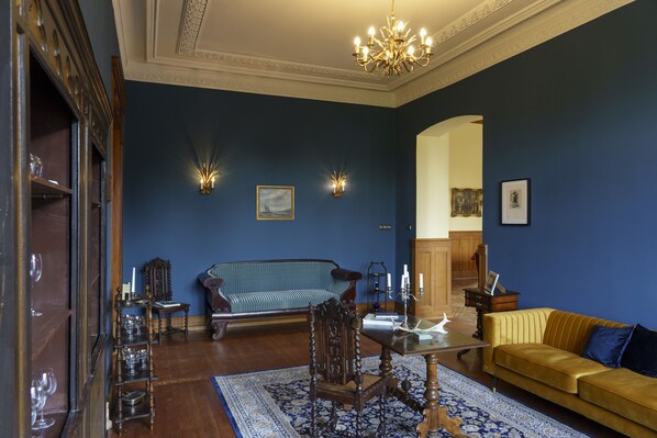 Une pièce avec un mur bleu, un plancher en bois, un canapé bleu, un canapé jaune, une chaise en bois, une table en verre et un lustre.
