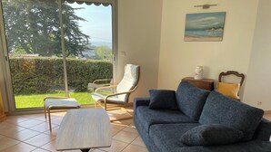 A living room with a sofa, a chair, a small table, and a view through a glass door to a garden.