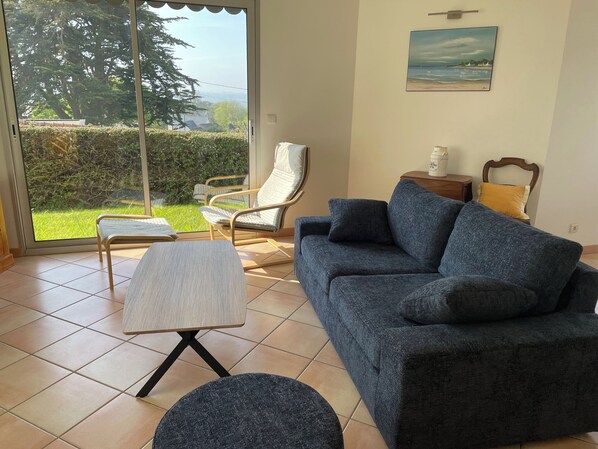 A living room with a sofa, a chair, a small table, and a view through a glass door to a garden.