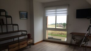 A room with a wooden floor, a desk with a chair, a window with blinds, and a TV mounted on the wall.
