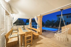 An outdoor patio with a wooden table and benches, a hanging chair, and a view of a swimming pool at dusk.