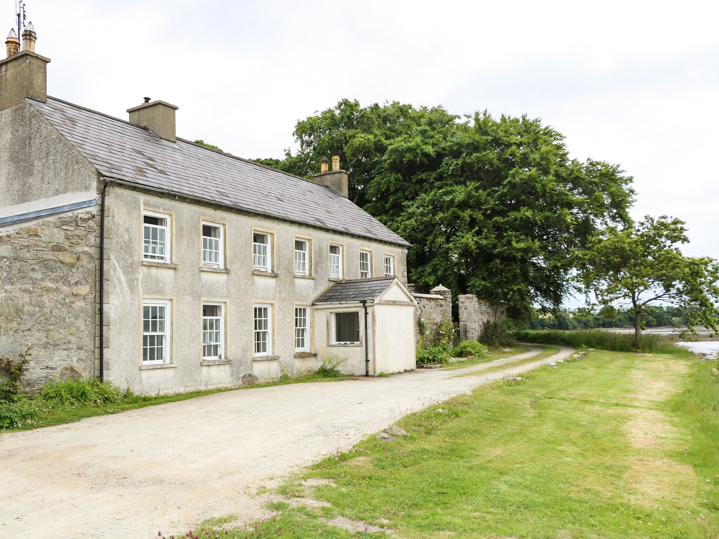 A stone building with multiple windows and a sloped roof, adjacent to a grassy area and a tree.