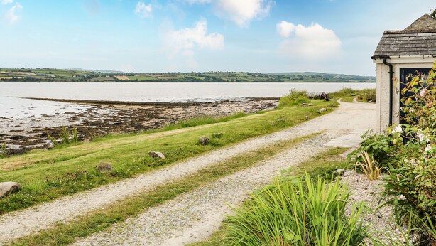 A coastal path leading to a small building with a sloped roof, surrounded by greenery and a view of the sea. - The Ferry House (Ramelton)