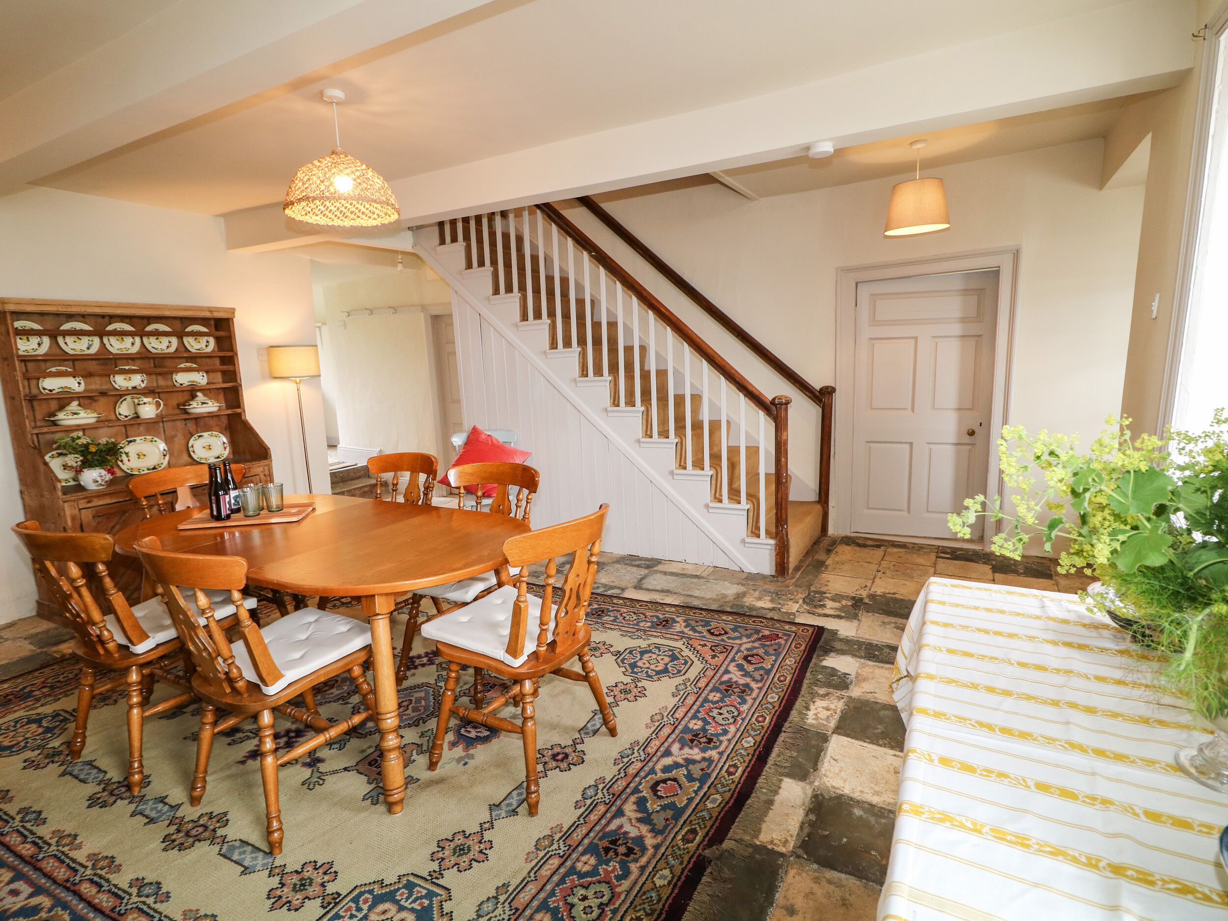 Un espace repas comprenant une table et des chaises en bois, un tapis à motifs, un escalier et une porte.