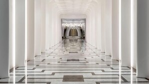 Un largo pasillo simétrico con columnas blancas y un suelo estampado, que conduce a una habitación con candelabro.