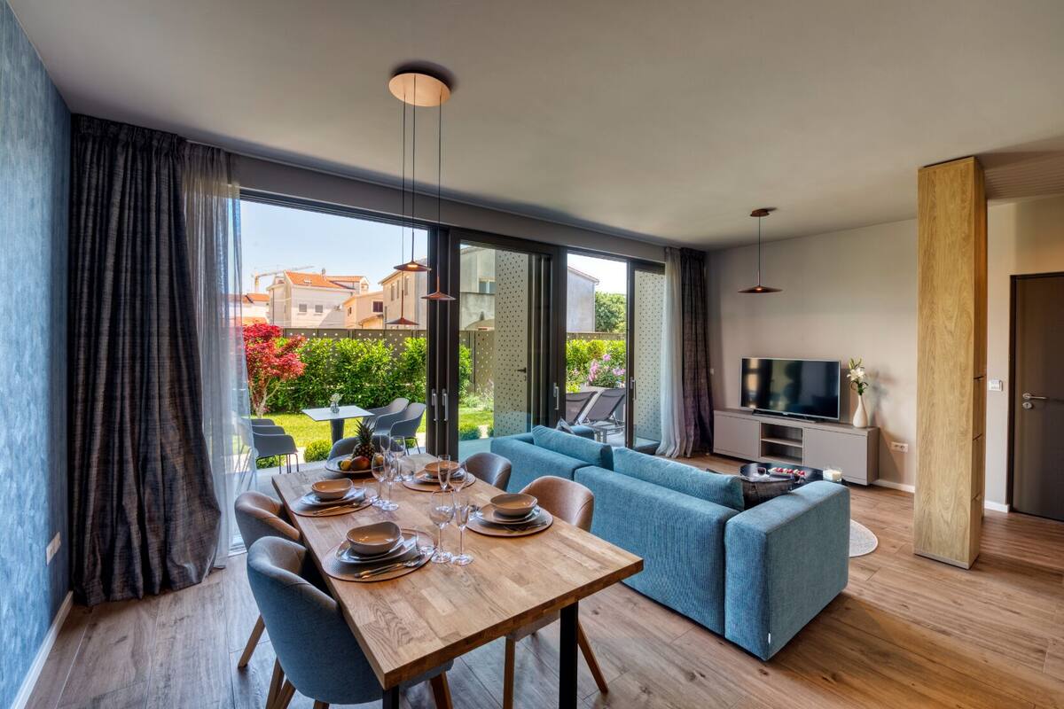 A modern living room with a wooden dining table set for four, a blue sofa, and a TV on a wooden cabinet.
