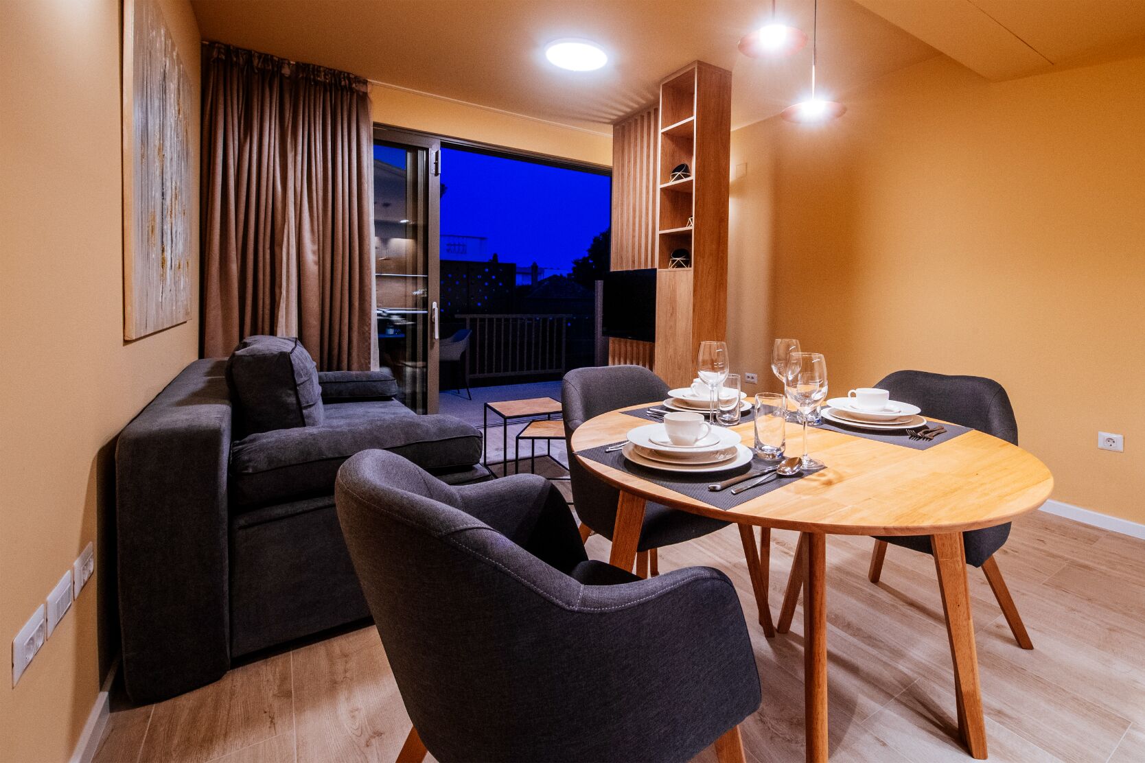 Ein modernes Wohnzimmer mit einer Couch, zwei Sesseln, einem Esstisch für zwei Personen und einer Glastür, die auf einen Balkon führt.