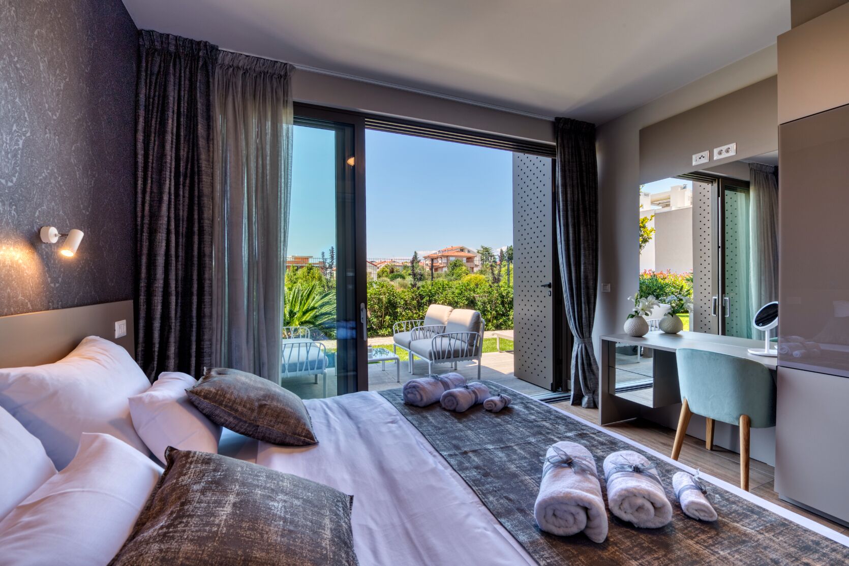 Habitación de hotel moderna con una cama grande, un escritorio con una silla, un pequeño balcón con vistas a zonas verdes y edificios, y un amplio ventanal.