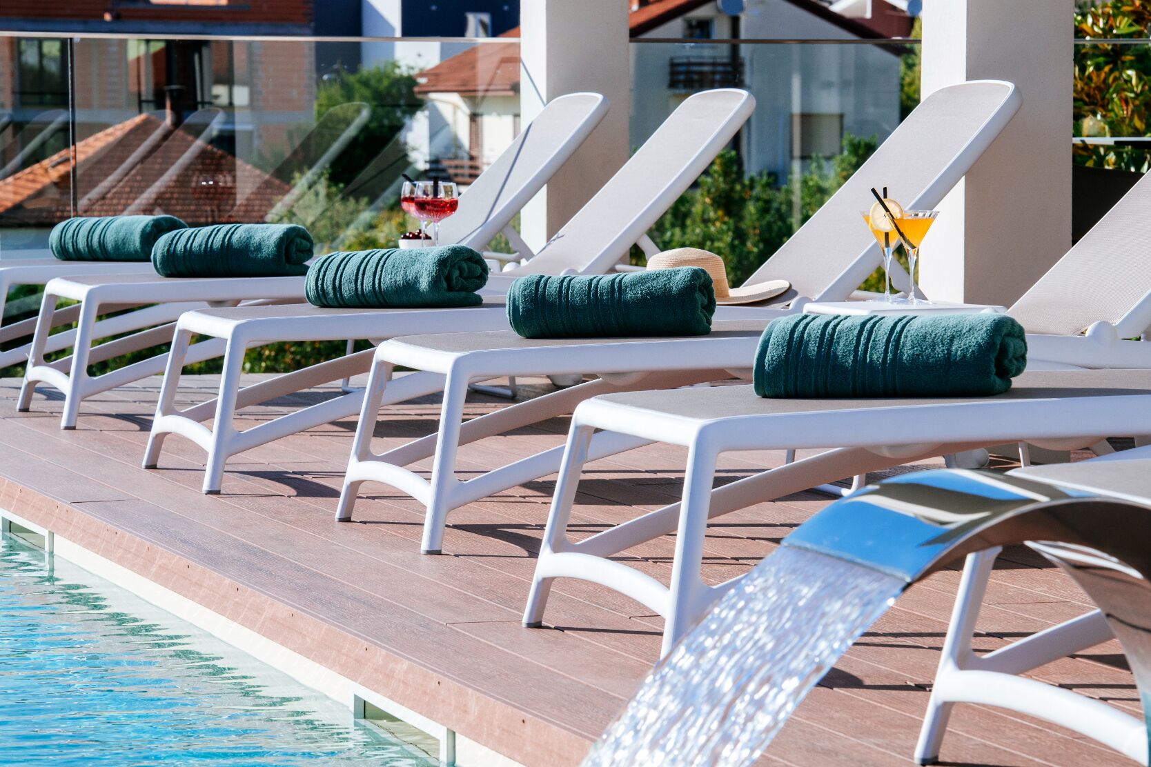 A poolside area with white lounge chairs and green towels, a glass of red drink, and a cocktail with a lemon twist.