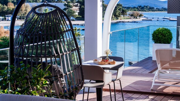 Un balcón con una silla de mimbre, una mesa con una bandeja de frutas y un florero, y vistas al mar y las montañas.
