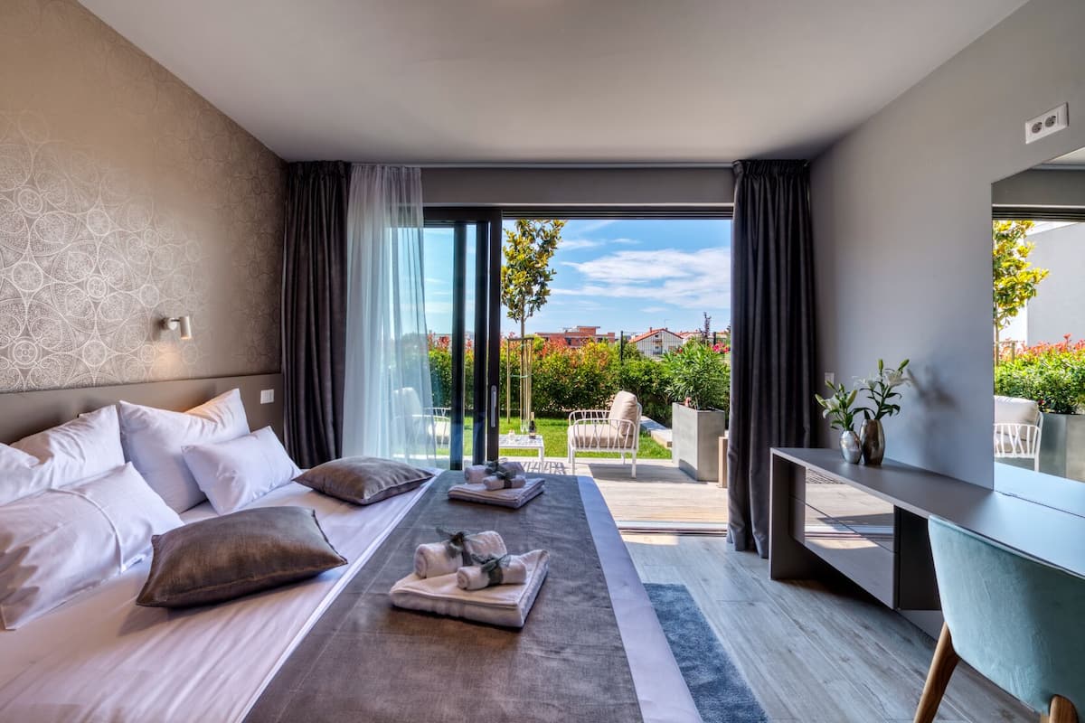 A modern hotel room with a large bed, a desk, and a view of a garden and buildings.