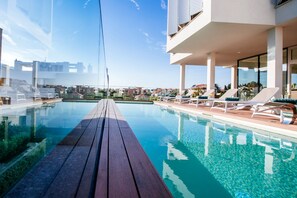 Un área de piscina moderna con una terraza de madera, sillas de descanso y vistas despejadas de los edificios circundantes.