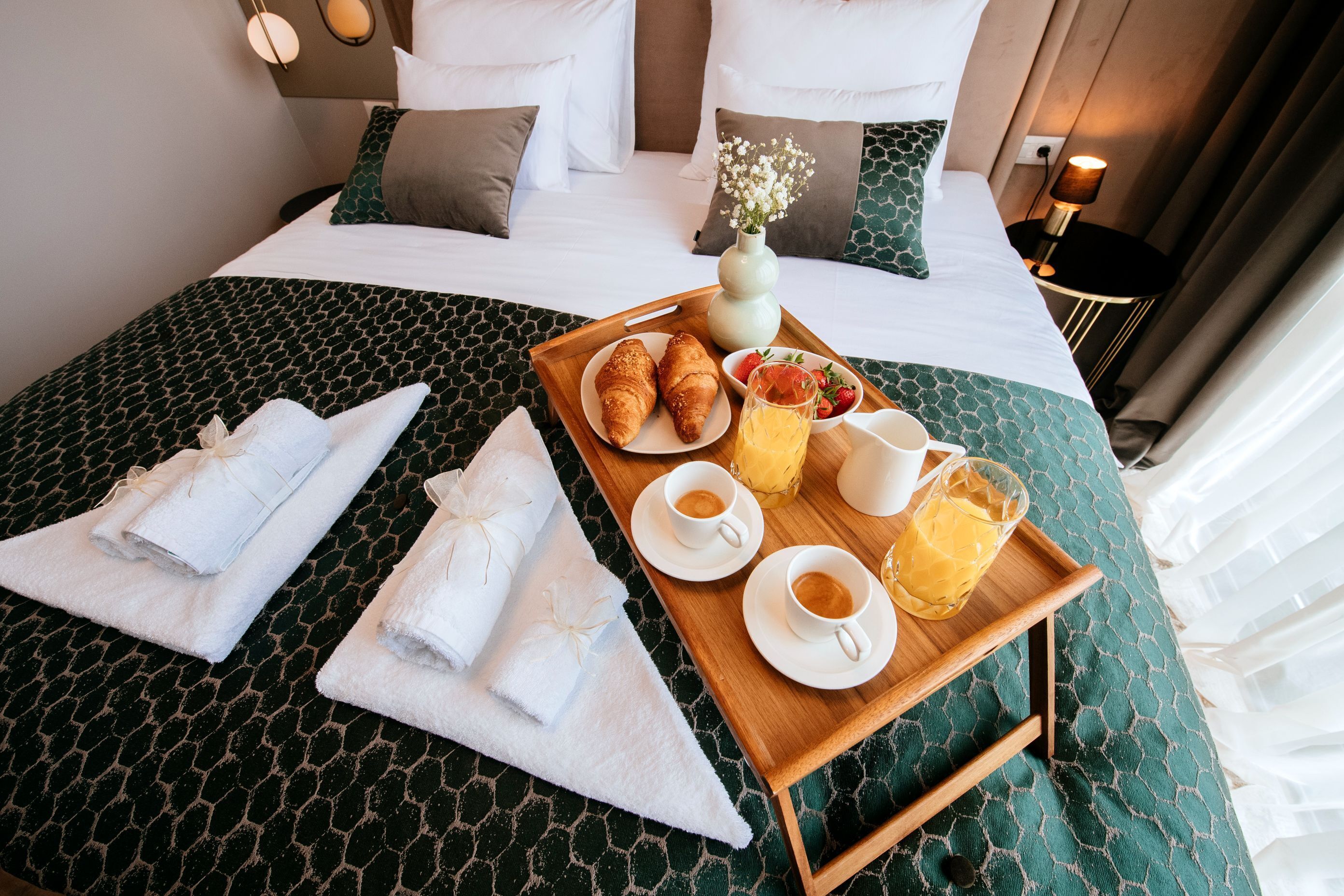 A neatly made bed with a green patterned bedspread, a tray with croissants, fruit, coffee, and a vase of flowers.