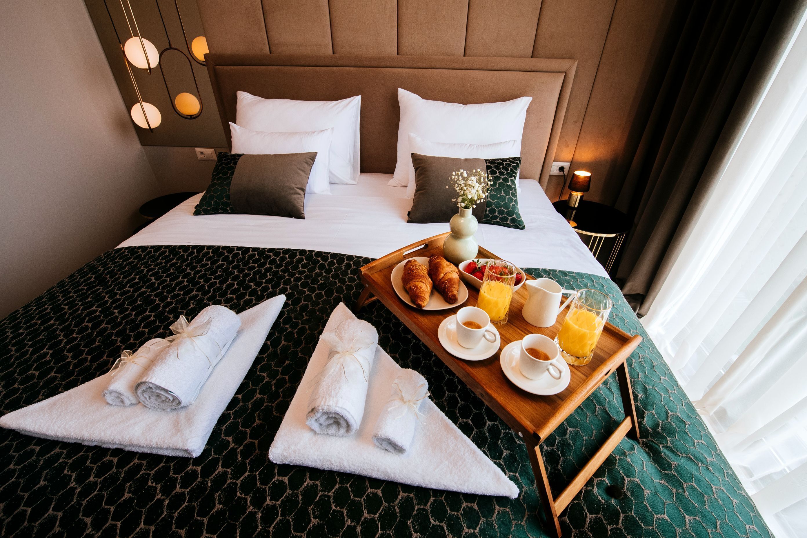 A neatly made bed with a tray of breakfast items, including croissants, fruit, and coffee, on a green patterned bedspread.