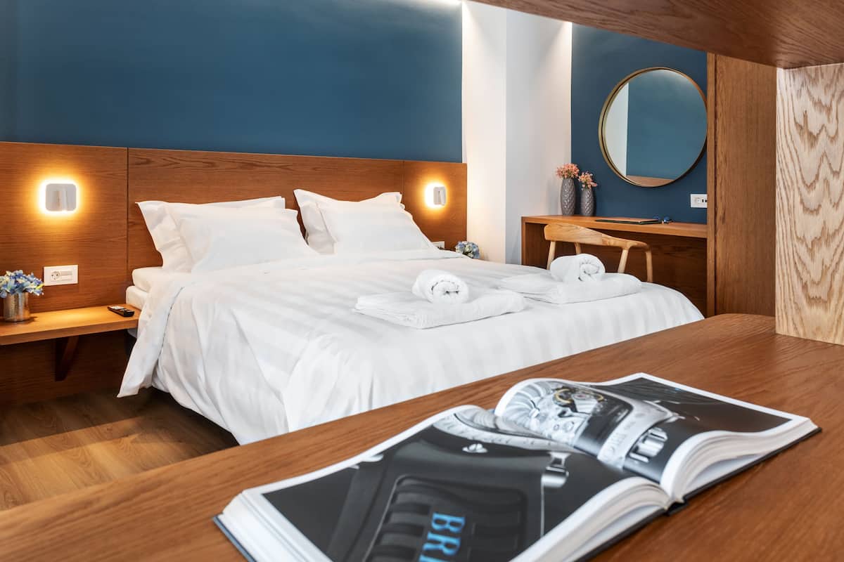 Una habitación de hotel moderna con cabecera de madera, una cama grande con ropa blanca, una mesita de noche con lámpara, otro mesita de madera y una revista sobre la mesa.