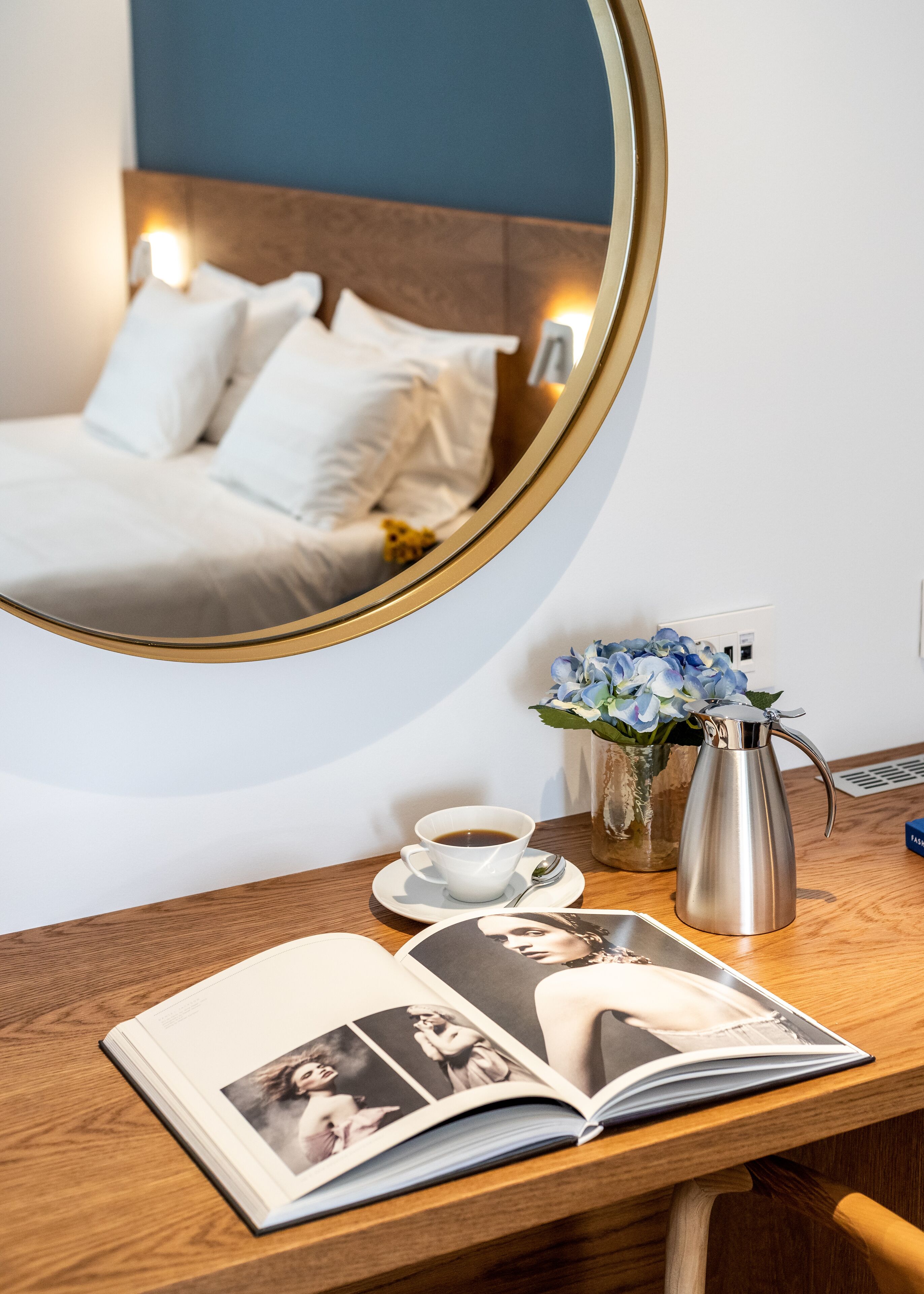 A hotel room with a bed, a bedside table with a coffee cup, a vase with flowers, and a magazine.