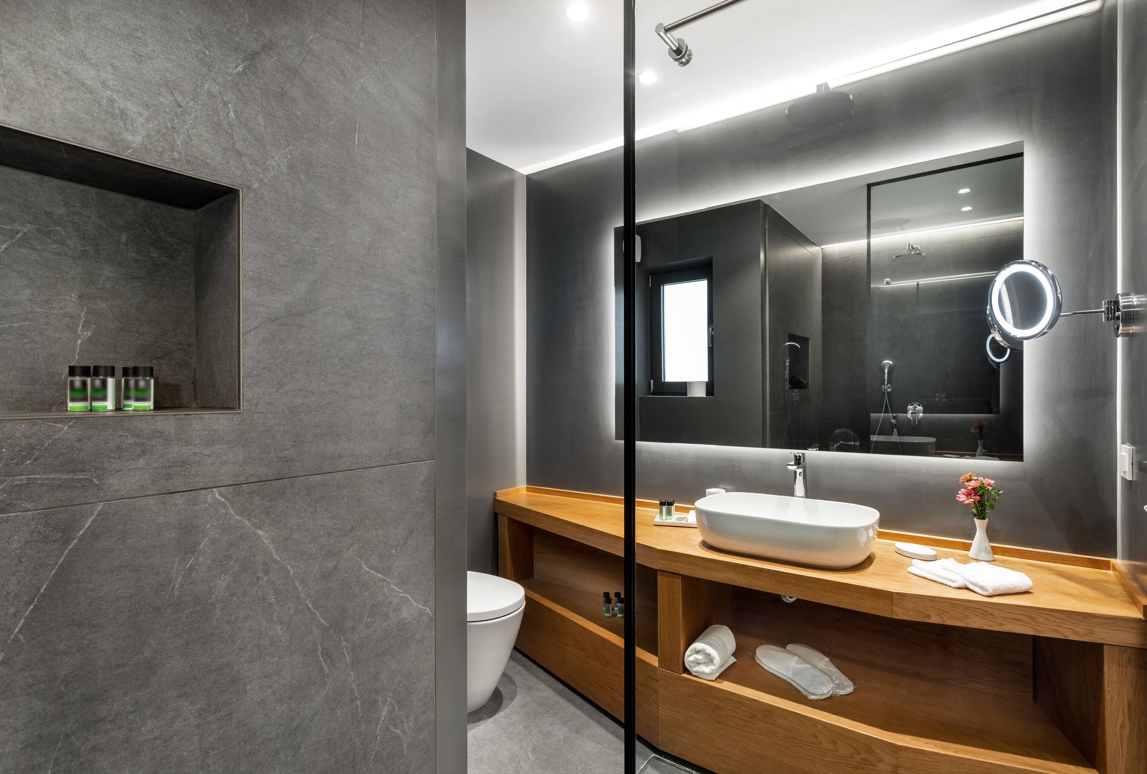 A modern bathroom with a large mirror, a wooden vanity, a built-in shelf with toiletries, and a glass shower enclosure.