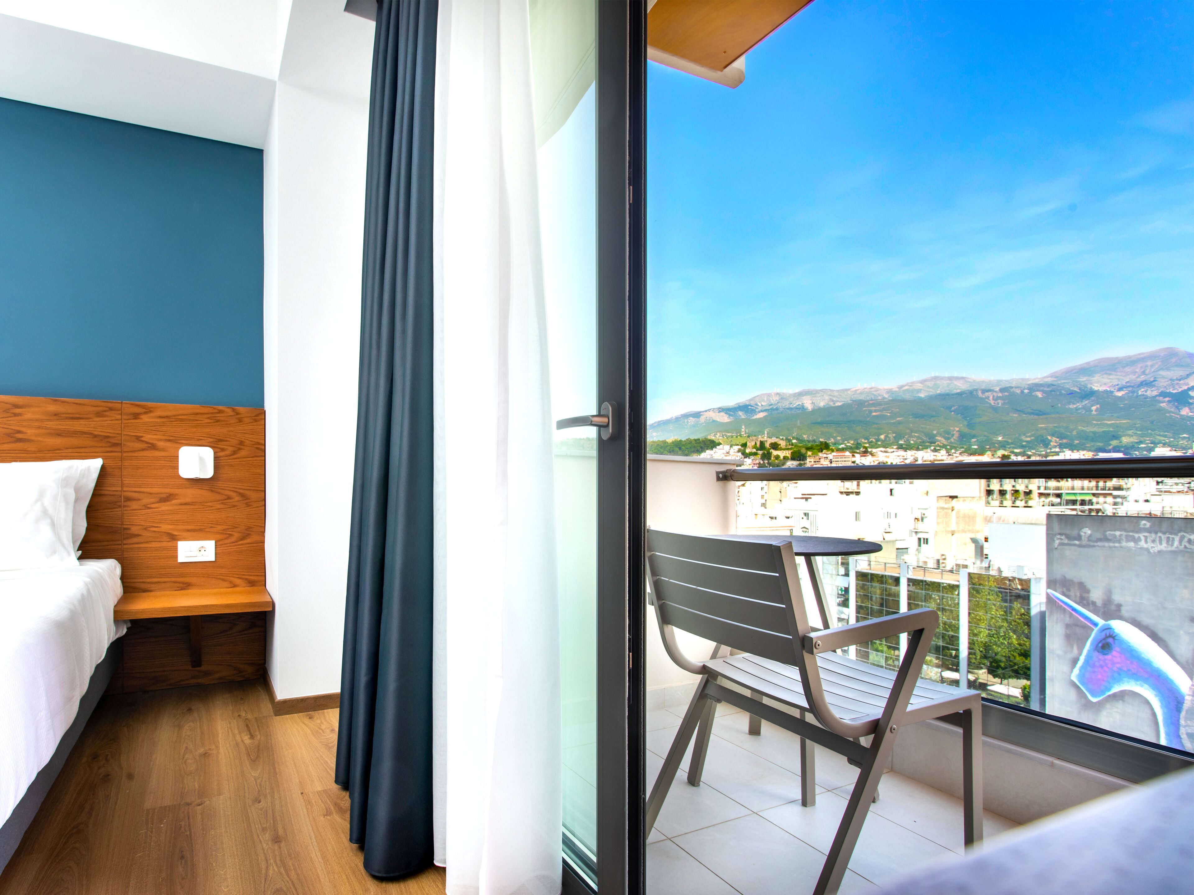 A hotel room with a bed, a wooden headboard, a balcony with a chair and table, and a view of a cityscape and mountains.