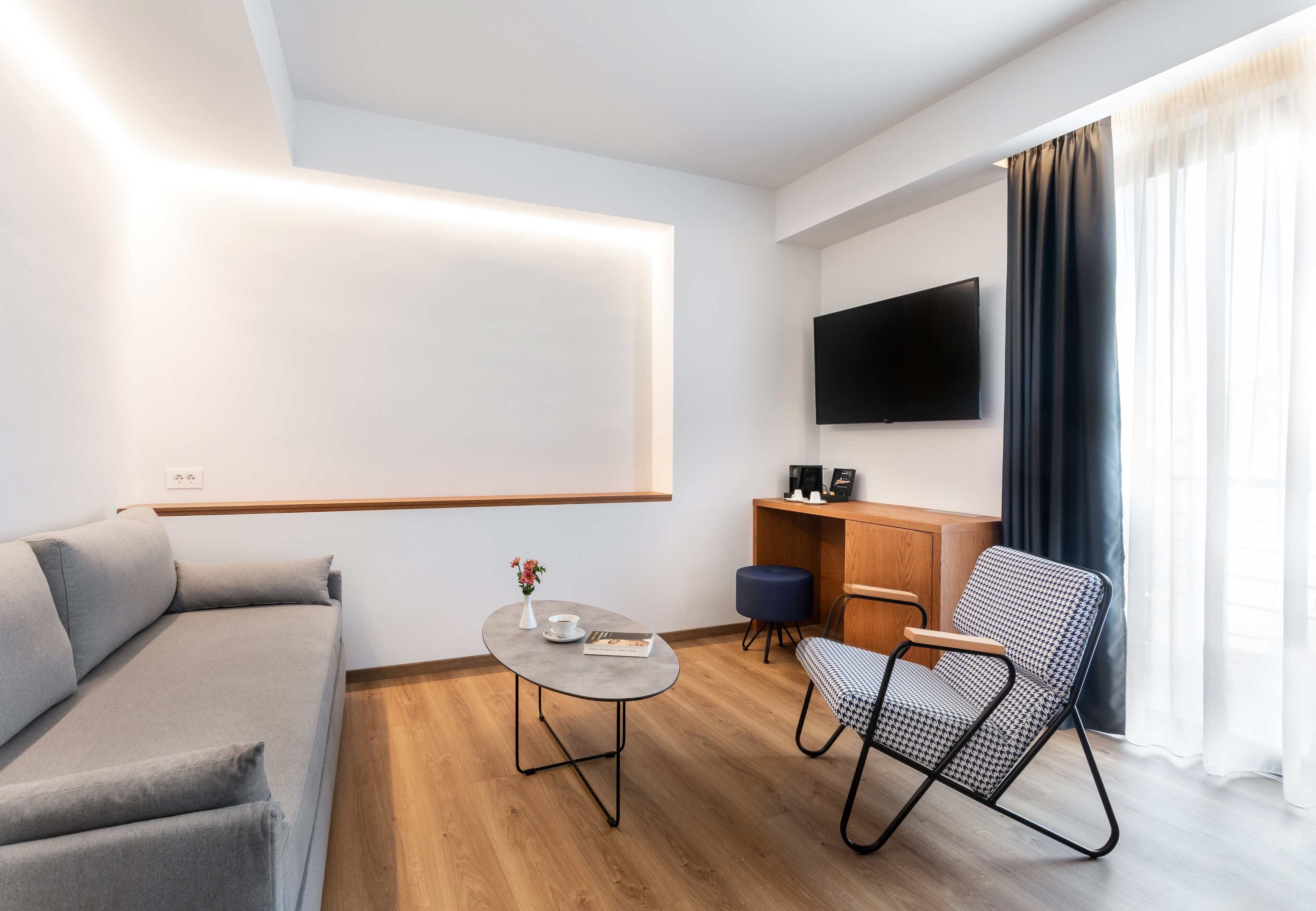A modern living room with a sofa, a small round table, a chair, a TV mounted on the wall, and a window with curtains.
