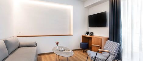 Una sala de estar moderna con un sofá, una mesita redonda pequeña, una silla, un televisor montado en la pared y una ventana con cortinas.