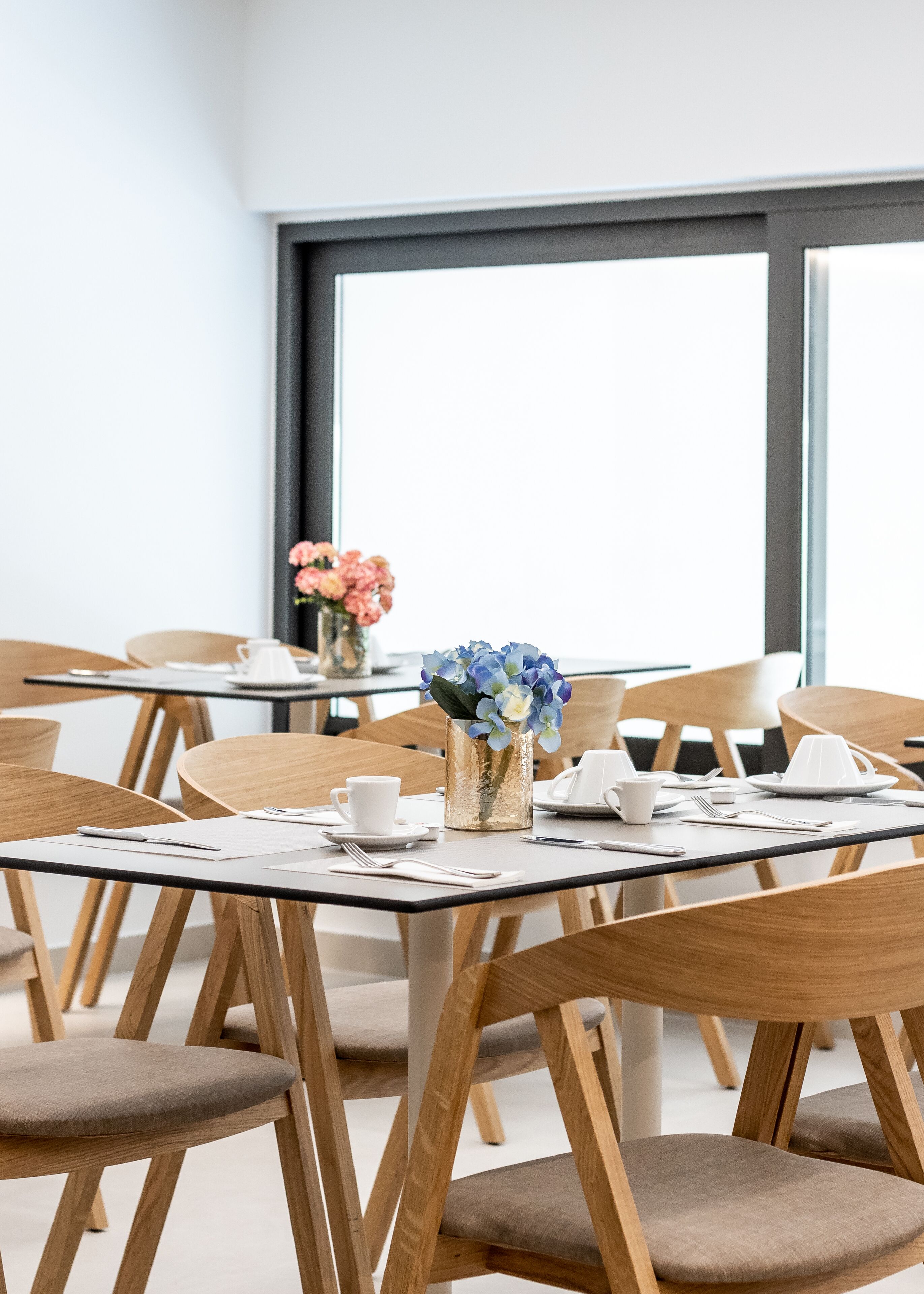 A modern dining area with wooden chairs and tables, a vase with flowers, and a large window in the background.