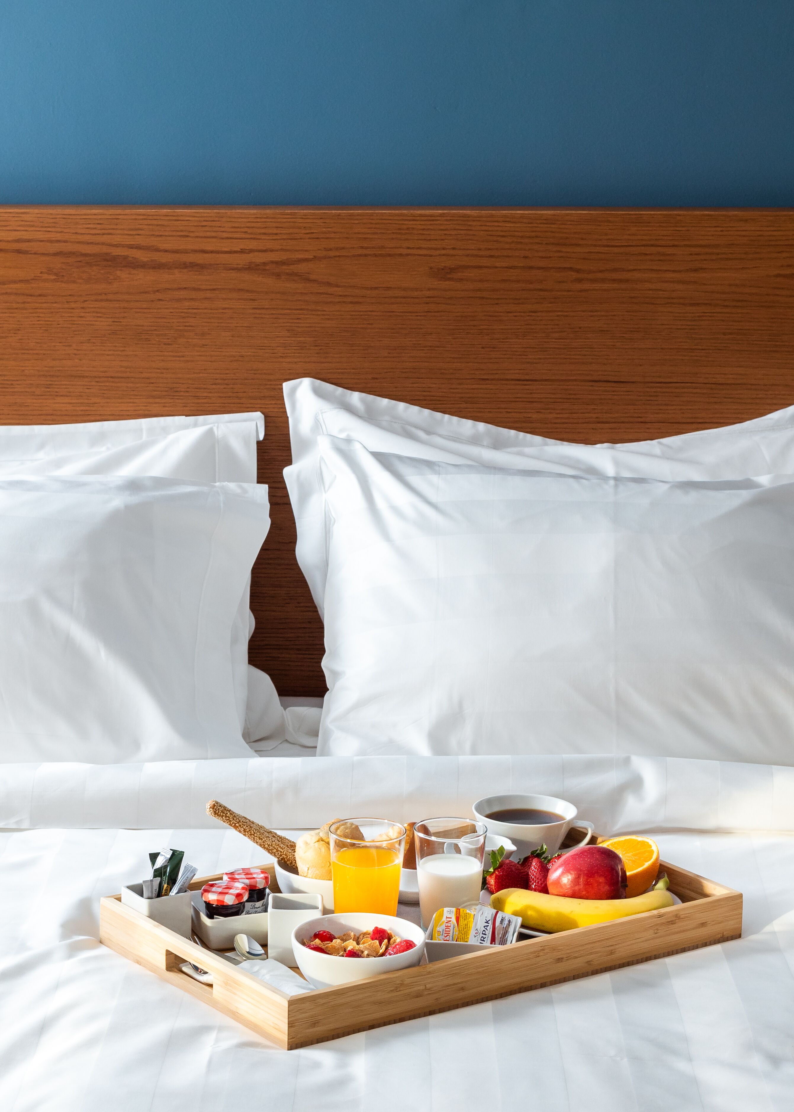 A neatly made bed with white linens, a wooden headboard, and a breakfast tray with cereal, fruits, juice, and coffee.