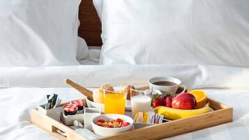 A neatly made bed with white linens, a wooden headboard, and a breakfast tray with cereal, fruits, juice, and coffee.