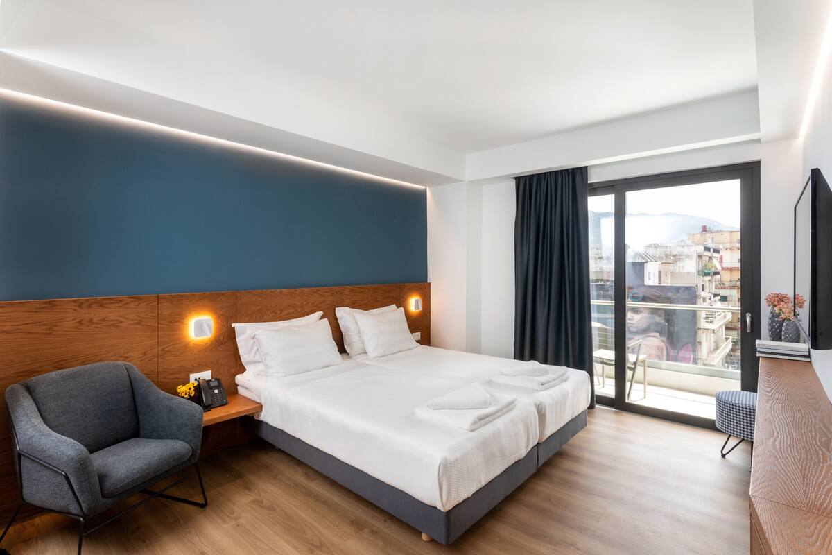 Una habitación de hotel moderna con una cama grande, una mesita de noche con lámpara, una silla y vistas a la ciudad a través de un ventanal amplio.
