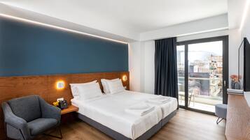 Una habitación de hotel moderna con una cama grande, una mesita de noche con lámpara, una silla y vistas a la ciudad a través de un ventanal amplio.