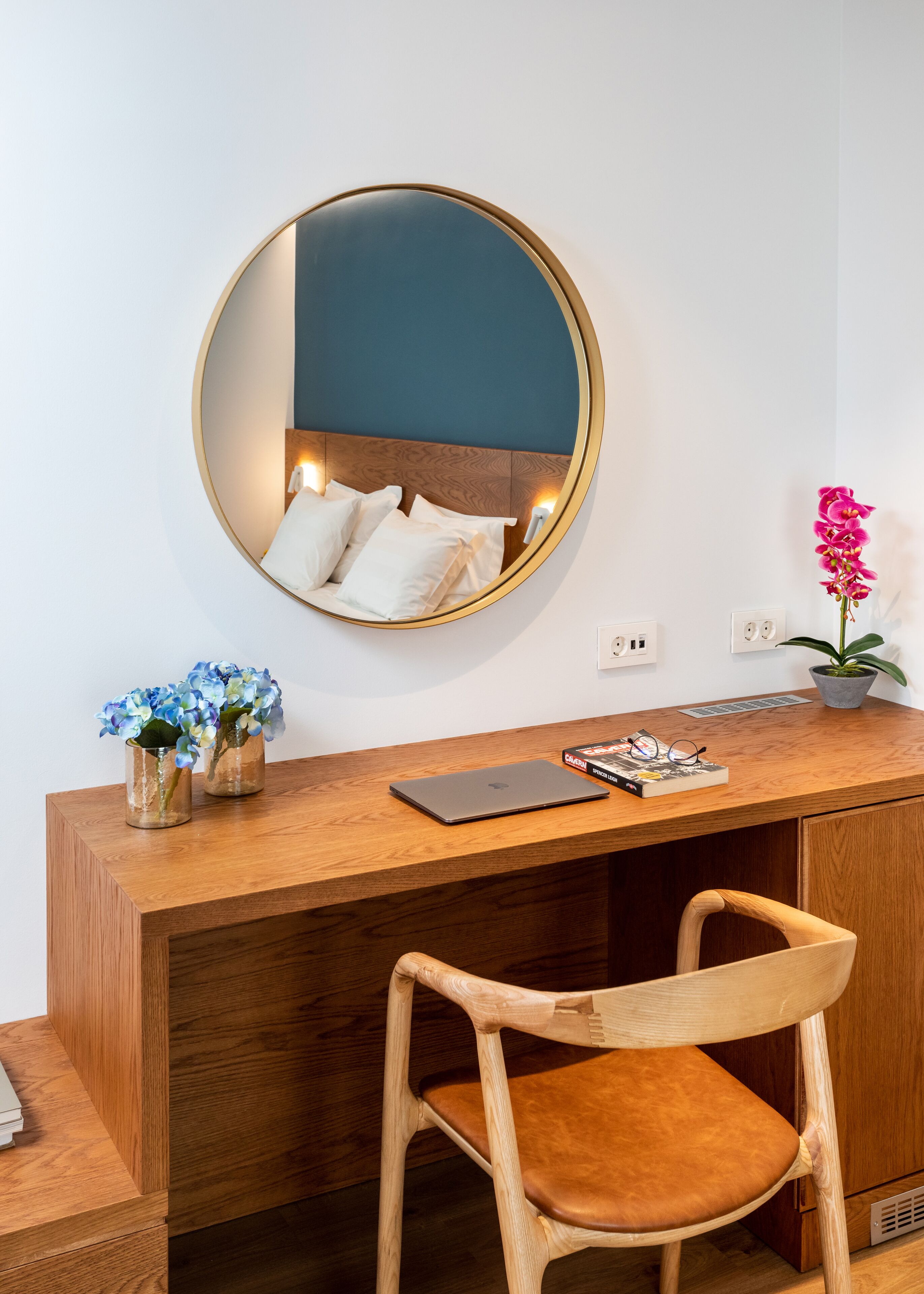 A wooden desk with a laptop, a mirror, and a vase with flowers.