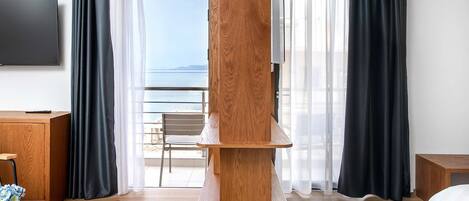 A hotel room with a wooden console table, a TV mounted on the wall, a bed, a chair, and a view of the sea through large windows.