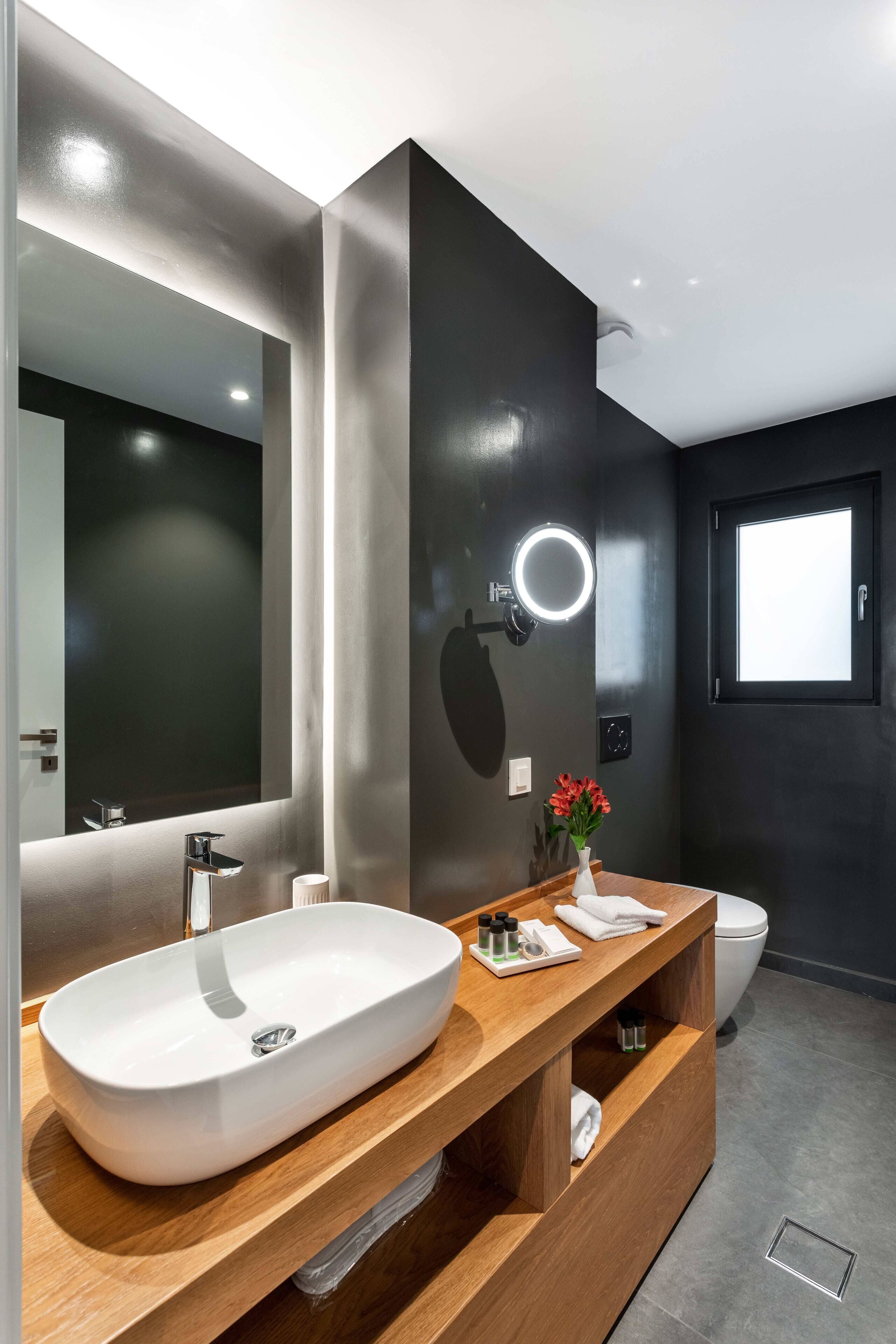 A modern bathroom with a large oval sink, a wooden vanity, a mirror with a round light, and a toilet.