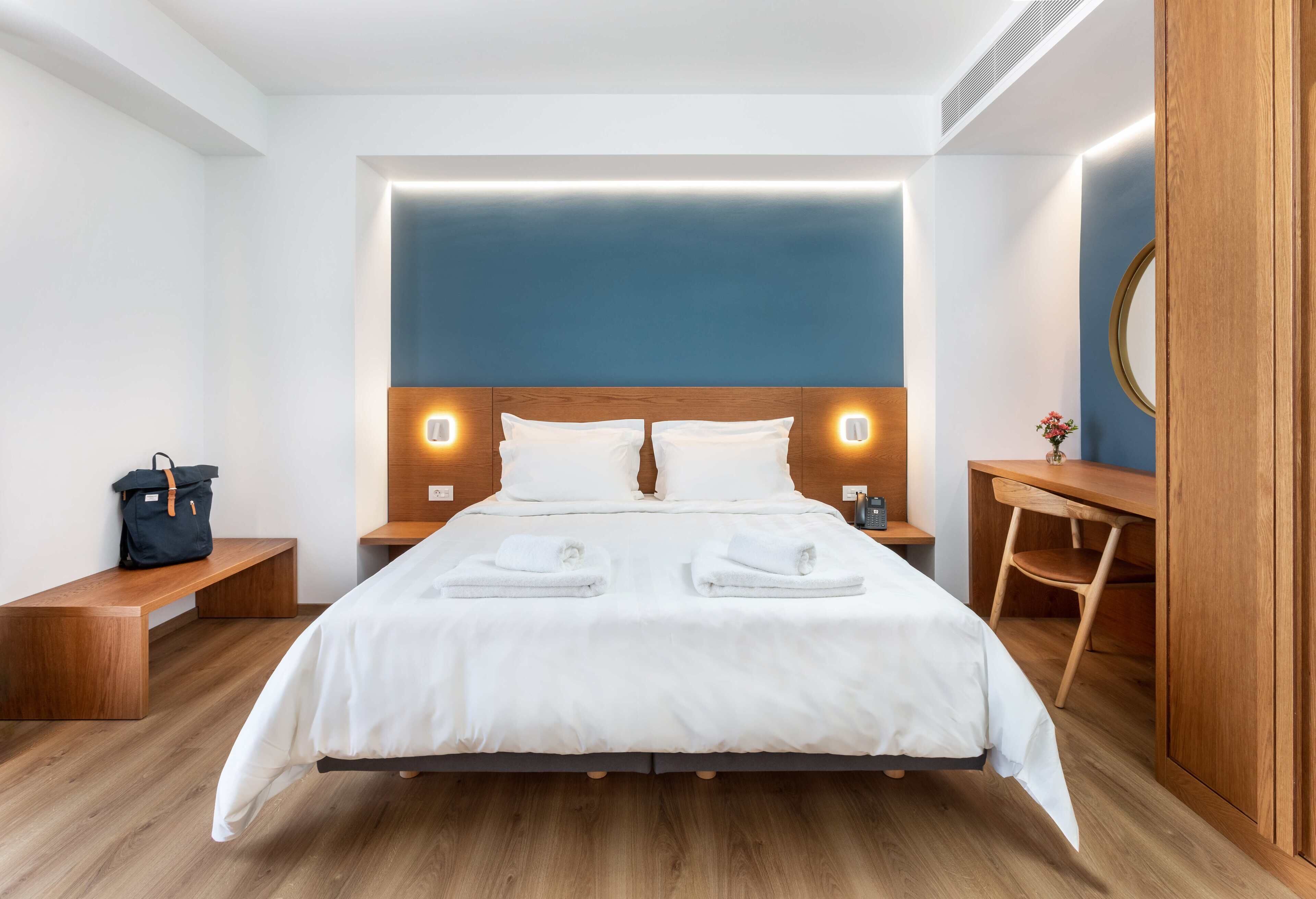 Een moderne hotelkamer met een groot bed, een houten hoofdbord, nachtkastjes, een bankje, een bureau en een spiegel.