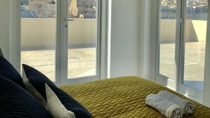 A room with two large arched windows, a bed with a quilted mattress, and a view of a cityscape with historical buildings.