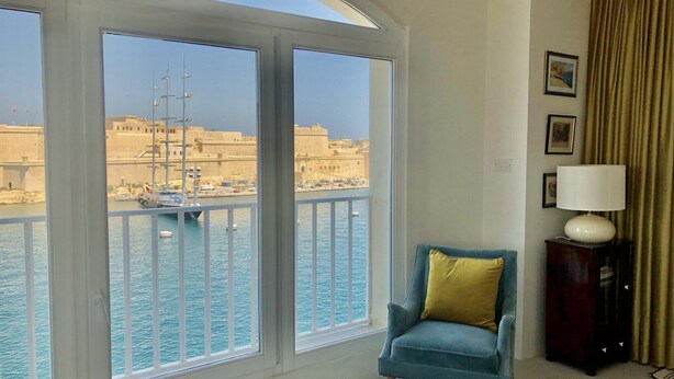 A room with a large window overlooking a harbor with boats and a building. There is a blue armchair with a yellow pillow, a lamp on a side table, and a curtain on the window.