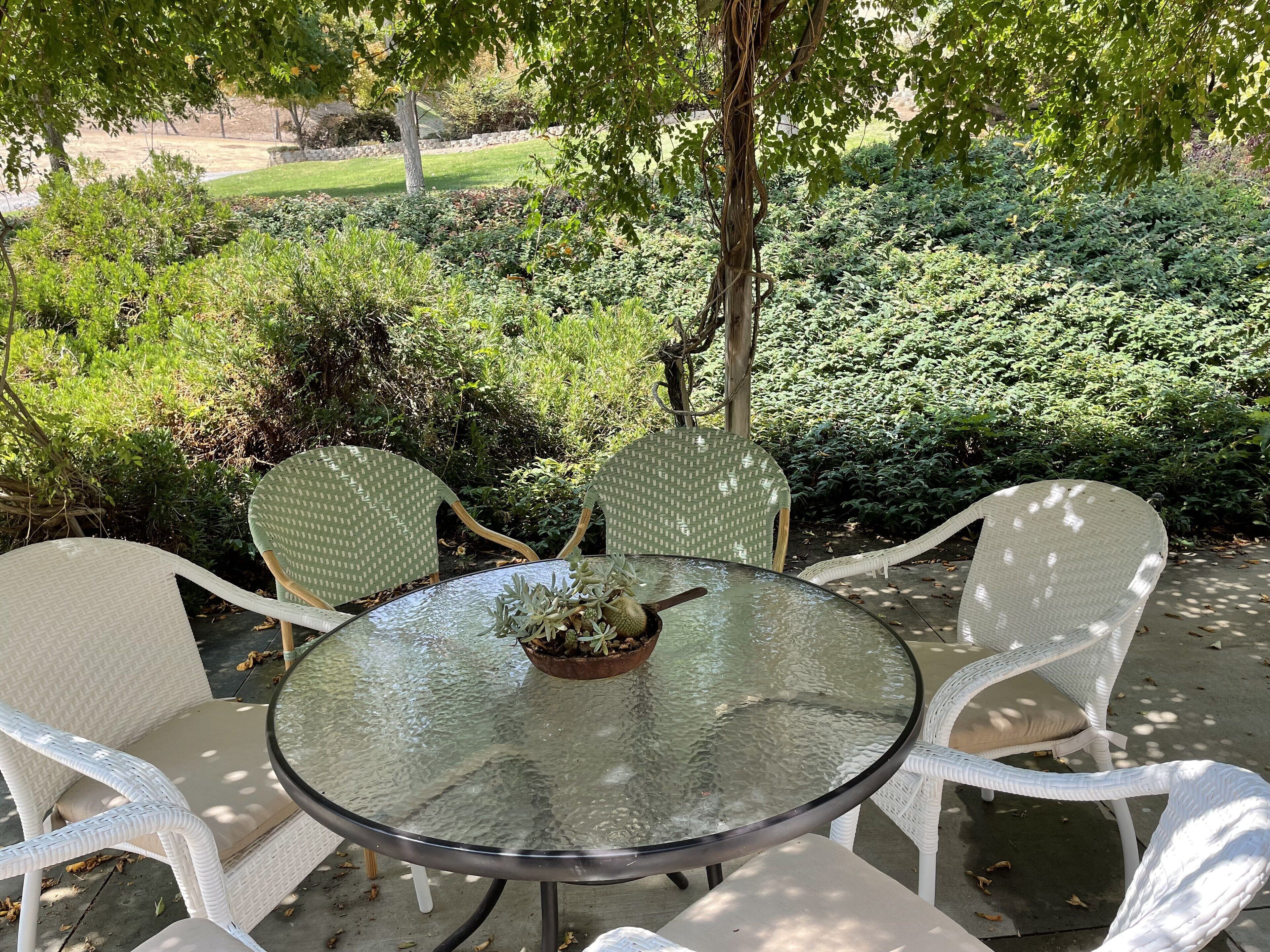 A round glass table with white wicker chairs, surrounded by lush greenery and a stone wall in the background.