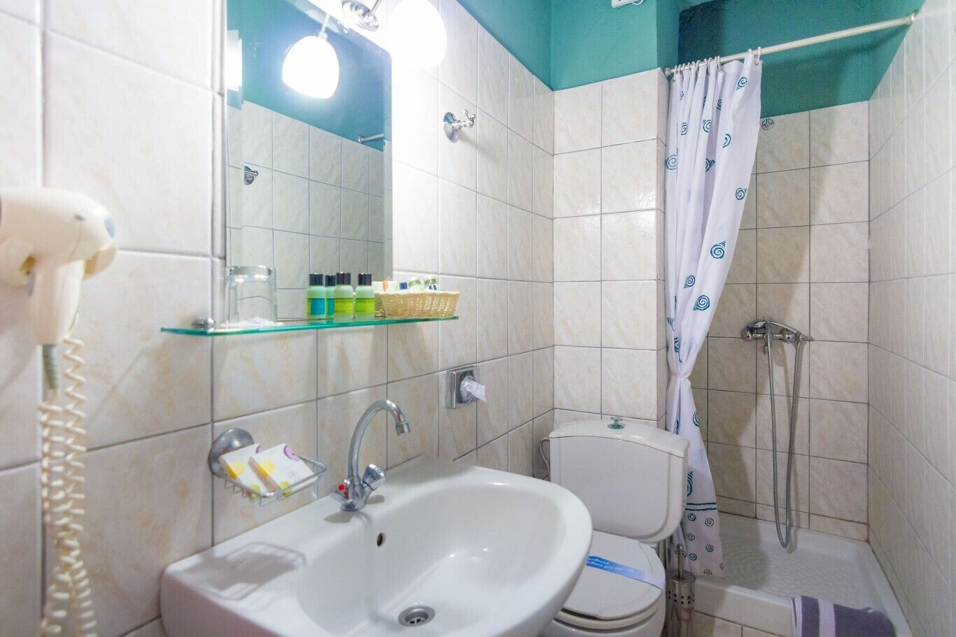 A compact bathroom with a sink, toilet, and shower. There is a glass shelf above the sink holding toiletries.