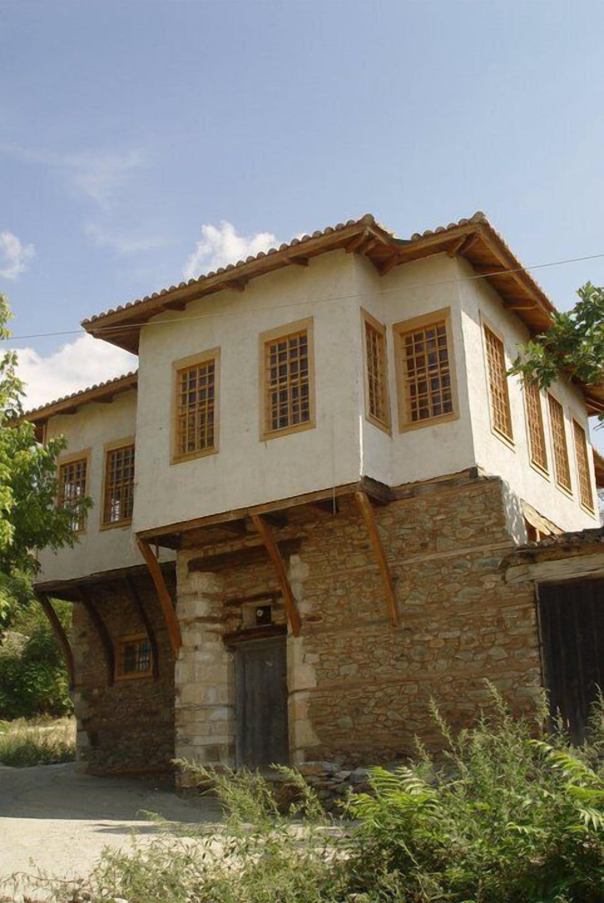 A two-story building with a stone base and a tiled roof. It has multiple windows with wooden frames and a central entrance with a door.