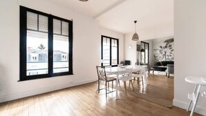 A spacious room with large windows, a dining area with a table and chairs, and a modern kitchen in the background.