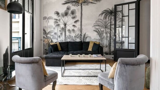 A modern living room with a black sofa, two grey armchairs, a coffee table, and a large window with a view of palm trees.