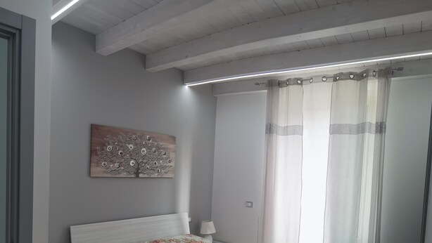 A bedroom with a bed, a nightstand, a lamp, a painting on the wall, and a window with curtains. - La Casa del Corso (Avola)