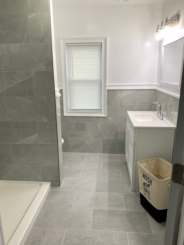 Un baño moderno con baldosas grises, una ventana con persianas, un lavamanos, un espejo y un cubo de basura con la etiqueta 'WASH AND DRY'.